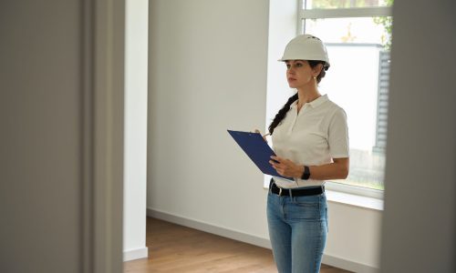 Focused adult caucasian female architect or designer writing in clipboard while looking at new empty modern comfortable townhouse